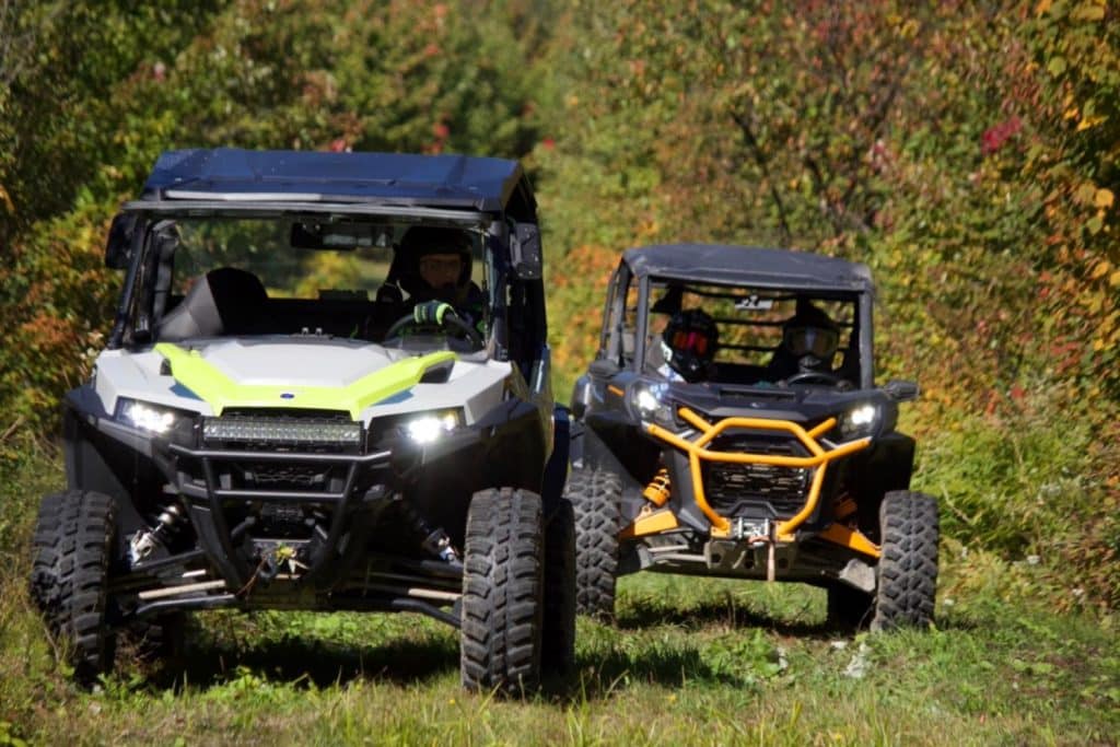 Deux véhicules côte à côte tout-terrain roulant sur un sentier forestier avec des conducteurs casqués au milieu d’une végétation colorée.