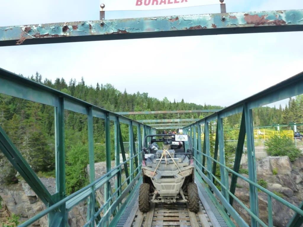VTT côte à côte traversant un pont métallique étroit au-dessus d’une rivière rocheuse, entouré d’une forêt dense sous un ciel dégagé.